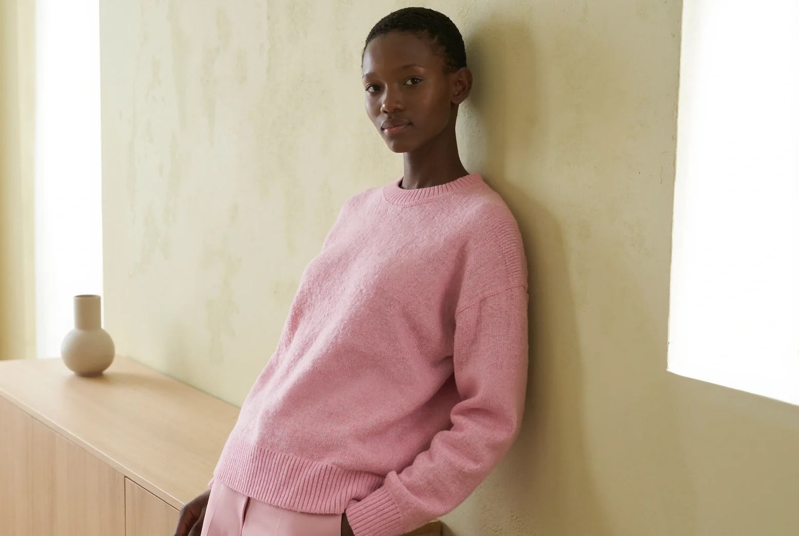 Model in a pink knit sweater leaning against a wall.